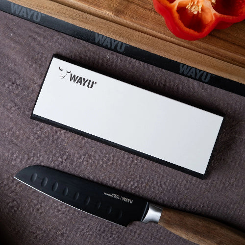 Cuchillo de cocina profesional Wayu con mango de madera, junto a su caja blanca sobre una superficie oscura, acompañado de una tabla de cortar y un pimiento rojo.