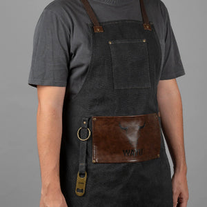 Persona usando un delantal de mezclilla gris oscuro con correas marrones y bolsillo frontal de cuero con el logo de Wayu.