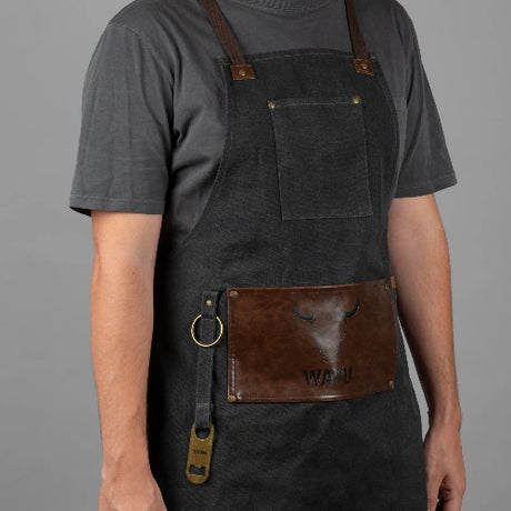 Persona usando un delantal de mezclilla gris oscuro con correas marrones y bolsillo frontal de cuero con el logo de Wayu.
