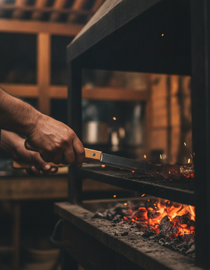 Manos sujetando pinzas parrillera mango madera sobre brasa parrilla chilena